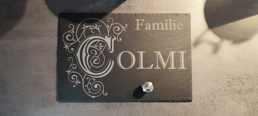 Rechteckige schwarze Schieferplatte mit heller Gravur „Familie COLMI“, großem verziertem Buchstaben C und silberner Klingel.