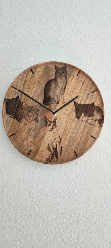 Runde Holzwanduhr mit schwarzen Zeigern und Strichmarkierungen. Auf dem Zifferblatt sind vier Katzenporträts eingraviert