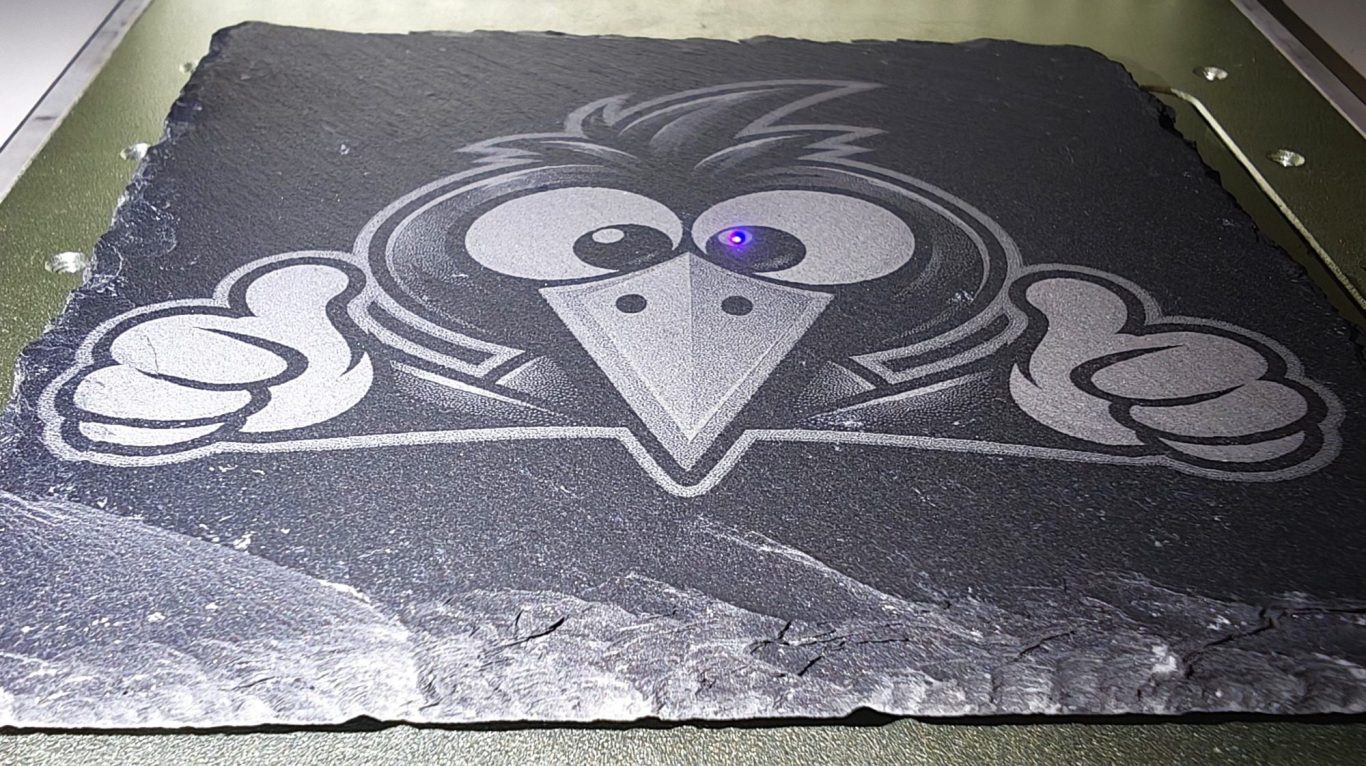 Schieferplatte mit humorvoller Vogel-Lasergravur Schwarze Schieferplatte mit heller Gravur eines lustigen Comicvogels mit großen Augen, spitzem Schnabel und zwei ausgestreckten Daumen.