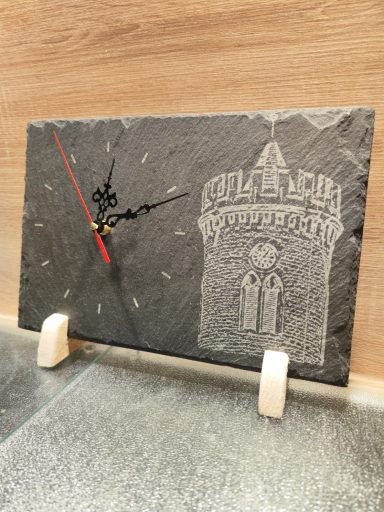 Rechteckige Schieferuhr mit heller Gravur eines historischen Steinturms, schwarzen Zierzeigern und rotem Sekundenzeiger auf zwei Holzfüßen.