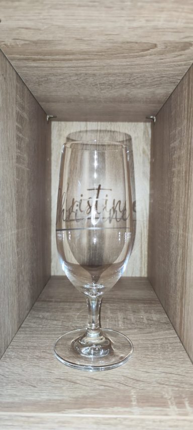 Klares Trinkglas mit Stiel und geschwungener Namensgravur „Christina“, stehend in einem hellen Holzregal.