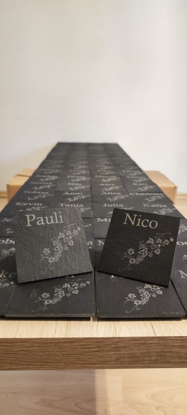 Zahlreiche schwarze Schieferuntersetzer mit hellen Namensgravuren und Weinreben. Vorne stehen zwei mit „Pauli“ und „Nico“.