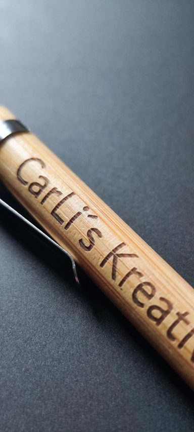 Nahaufnahme eines Bambus-Kugelschreibers mit dunkler Gravur „CarLi’s KreativDeko“ und silberfarbenem Metallclip auf schwarzem Hintergrund.
