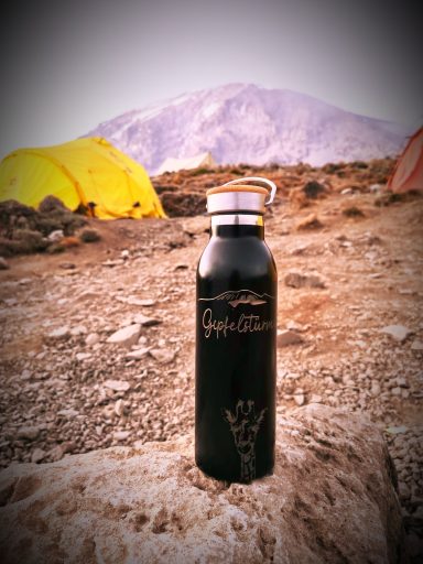 Schwarze Edelstahlflasche auf einem Felsen vor Bergkulisse und Zelten, graviert mit „Gipfelstürmer“, Bergen und Tierkopf.