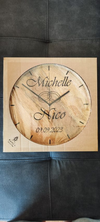Runde Holzuhr mit schwarzen Zeigern, den gravierten Namen „Michelle“ und „Nico“, einem Ornament und dem Datum „09.09.2023“.